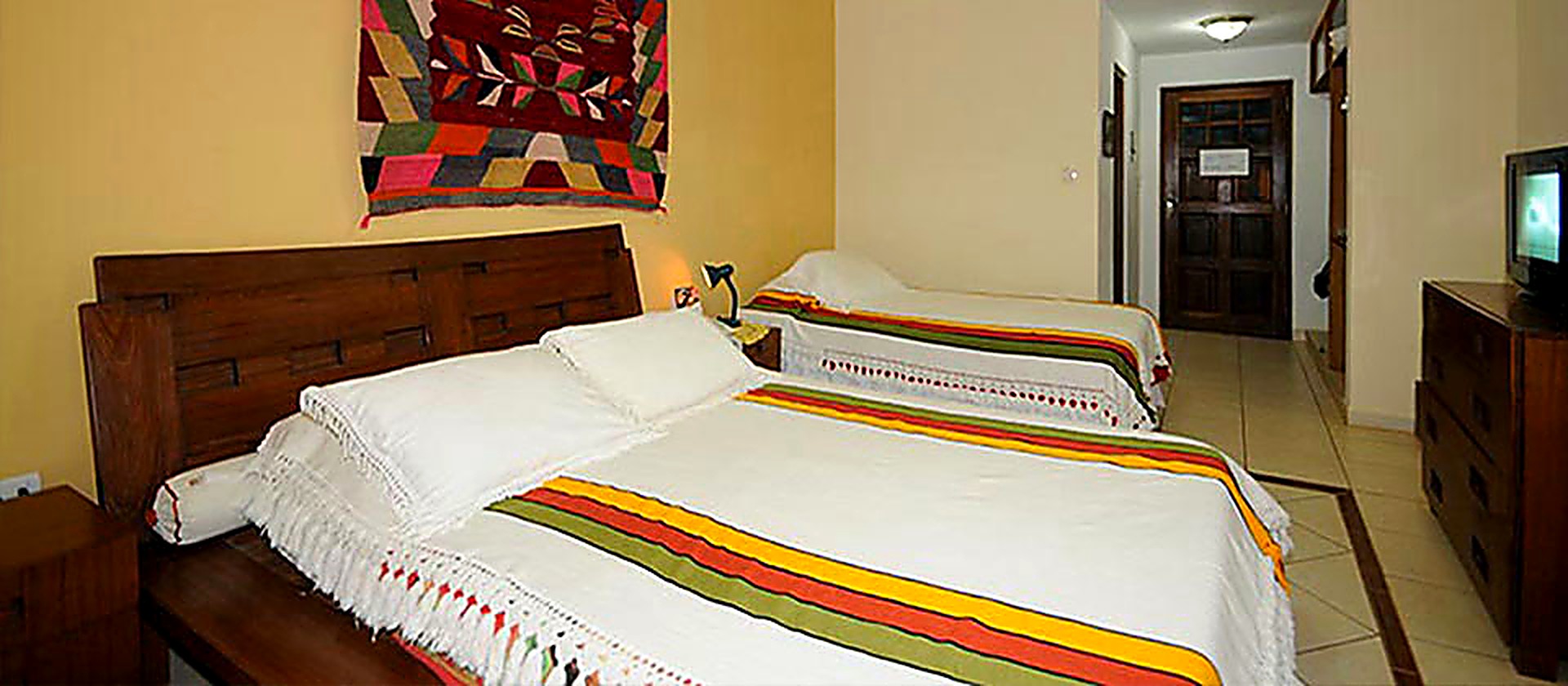 Habitación con cama matrimonial, cama individual y textil chiquitano en la pared